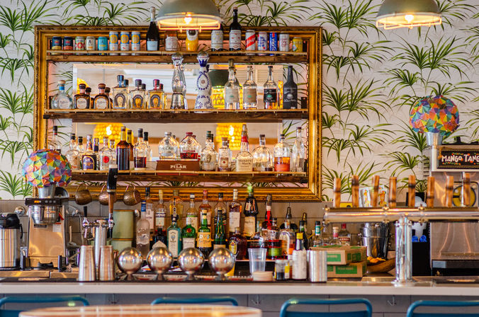 Vibrant tropical-themed cocktail bar interior with framed mirror backbar stacked with liquor bottles, colorful mosaic lamps, hanging pendant lights and multiple draft taps at a white counter.