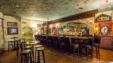 Cozy vintage pub interior with curved wooden bar, leather barstools, brick arch and mirrored shelves stocked with liquor, decorative tin ceiling and warm wood floors