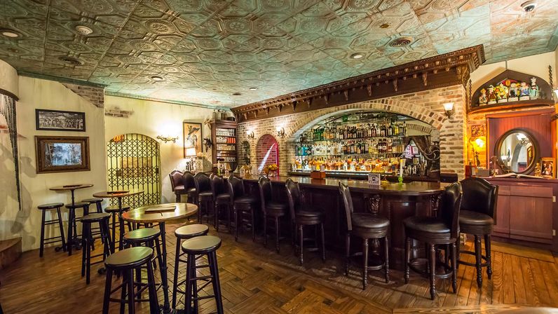 Cozy vintage pub interior with curved wooden bar, leather barstools, brick arch and mirrored shelves stocked with liquor, decorative tin ceiling and warm wood floors