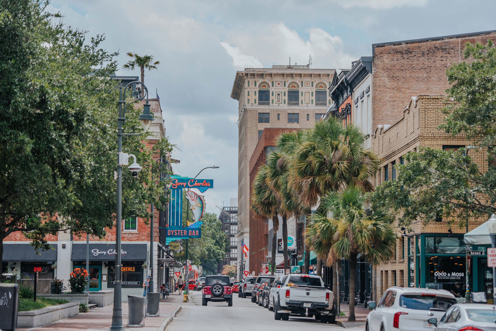 Downtown coastal street with palm trees, historic brick buildings, parked cars and a colorful neon fish oyster bar sign under a cloudy sky