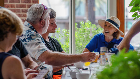Friends enjoying a casual brunch at a sunlit window-side café table — a man passes a drink to a laughing woman in a wide-brim hat with greenery visible outside.