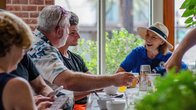 Friends enjoying a casual brunch at a sunlit window-side café table — a man passes a drink to a laughing woman in a wide-brim hat with greenery visible outside.