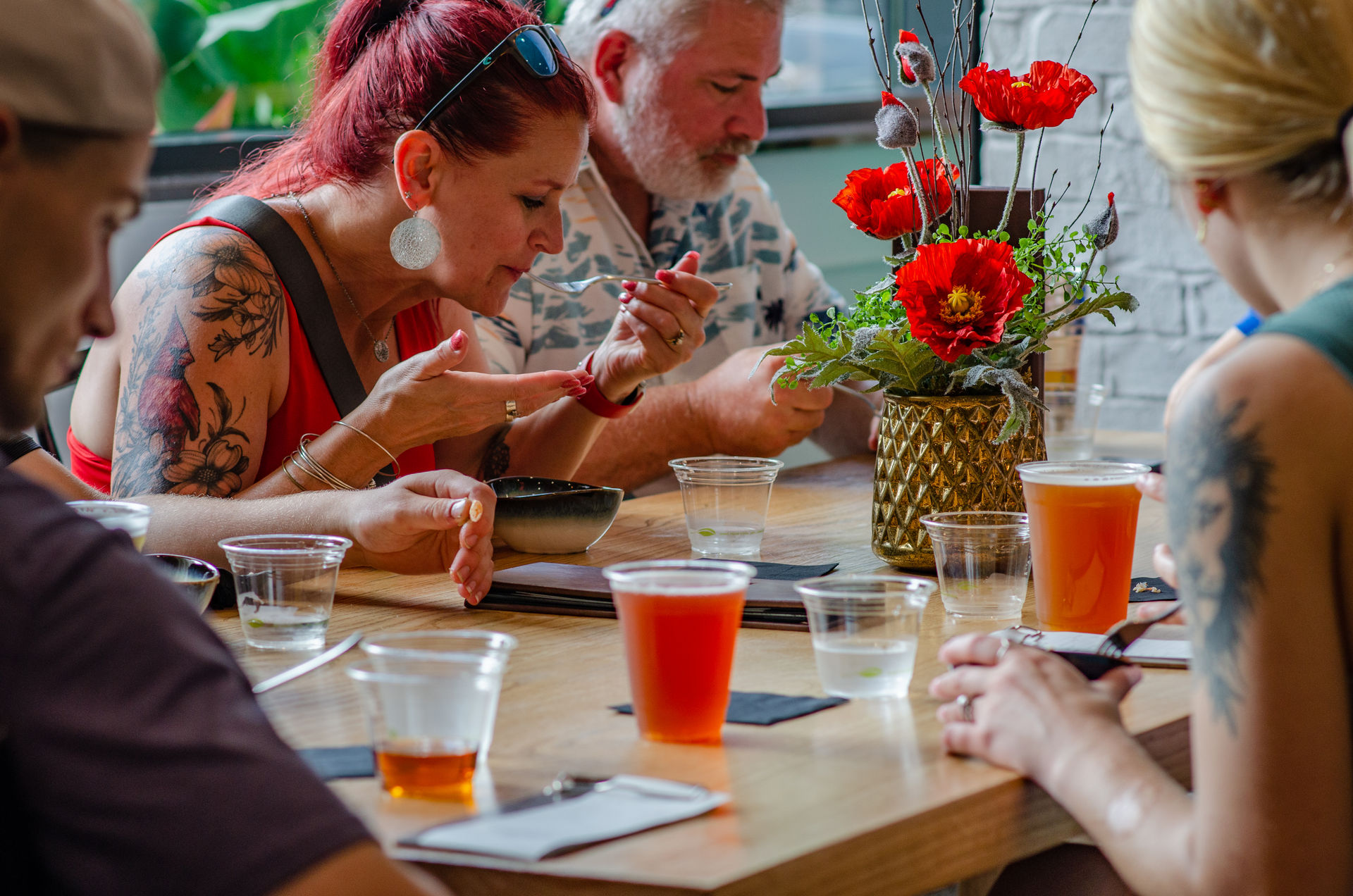 Group at a wooden communal table enjoying craft beers and bowls — a red-haired woman tasting food with a spoon, clear plastic cups and a gold vase with bright red flowers as the centerpiece.