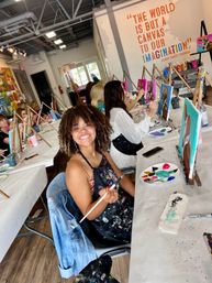 Smiling woman in a lively indoor group painting class at an art studio, holding a paintbrush among easels, colorful canvases, and a wall quote