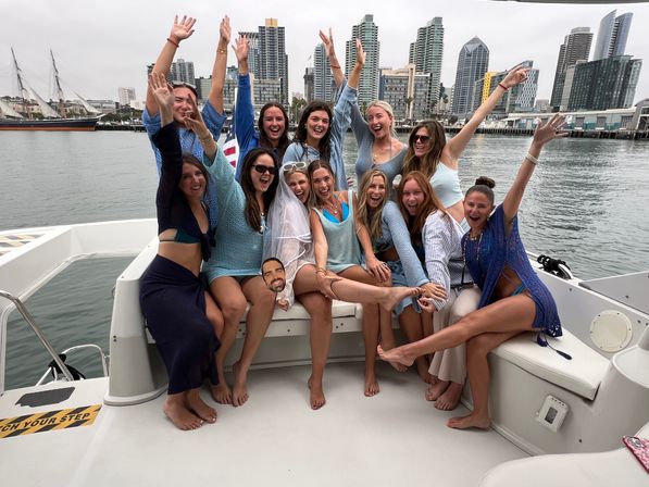 Smiling group of friends on a boat celebrating a bachelorette party, wearing summer outfits and a veil, cheering with arms raised in front of a city harbor skyline.