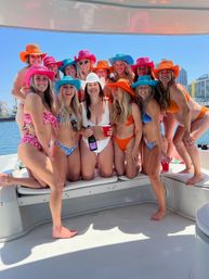 Boat party with a group of smiling women in bikinis wearing colorful cowboy hats (pink, orange, turquoise, white) on a sunny city harbor; one holds a bottle and red cup against a skyline backdrop.