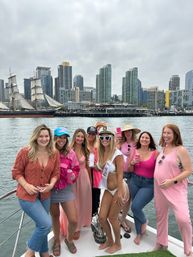 Group of women enjoying a bachelorette party on a boat at a city waterfront — bride in a white bikini, sash and captain hat with heart sunglasses, friends in pink outfits holding drinks, tall ship and high‑rise skyline under a cloudy sky.