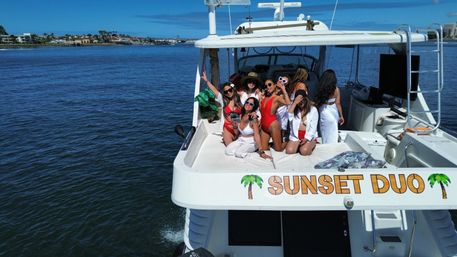 Group of women in red and white swimsuits posing and laughing on the upper deck of a white yacht, sunlit blue water and a palm-lined coastal skyline in the background.