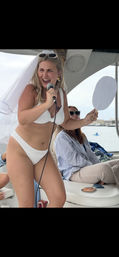 Laughing bride-to-be in a white bikini and veil sings into a microphone aboard a boat at a bachelorette party, holding a handheld fan while friends relax and a harbor appears in the background.