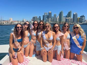 Eight women in bikinis wearing bachelorette sashes on a sunny boat in San Diego harbor with city skyline and bright blue sky