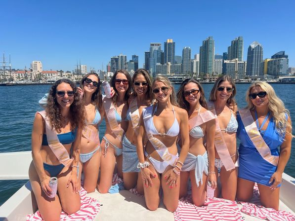 Eight women in bikinis wearing bachelorette sashes on a sunny boat in San Diego harbor with city skyline and bright blue sky