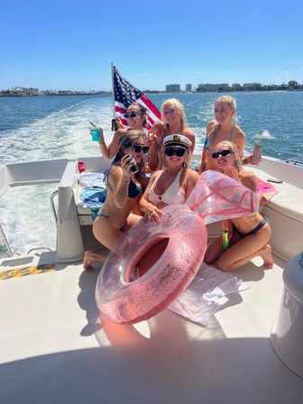 Six women in bikinis partying on a motorboat in a sunny Southern California bay with an American flag, captain’s hat and a sparkly pink inflatable donut