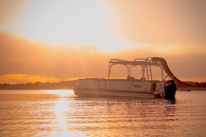 Pontoon boat with waterslide anchored on a calm lake at golden-hour sunset, warm orange sky reflecting on rippled water.