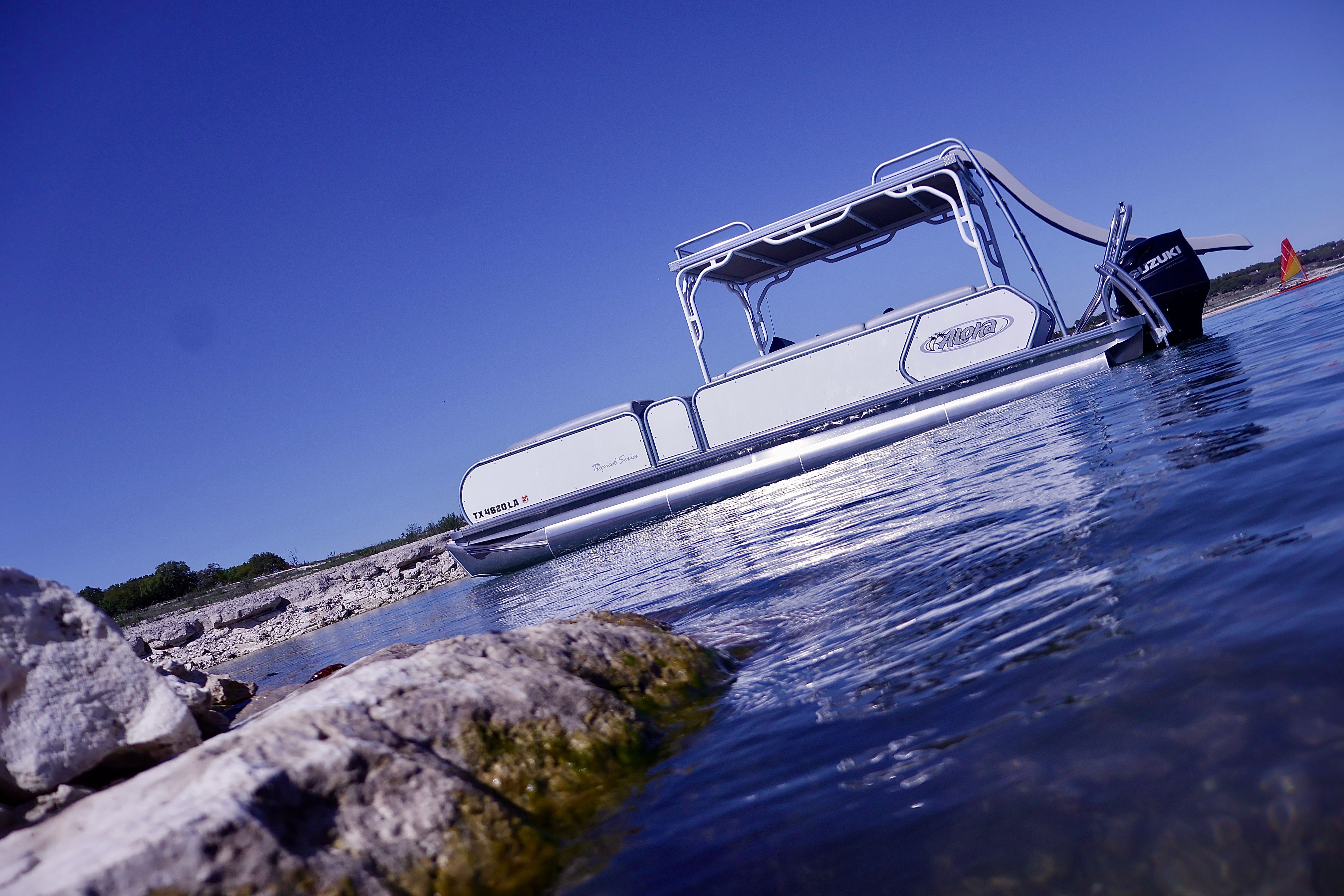 White pontoon boat moored near a rocky lakeshore with calm blue water reflecting sunlight and a distant red sailboat under a clear sky