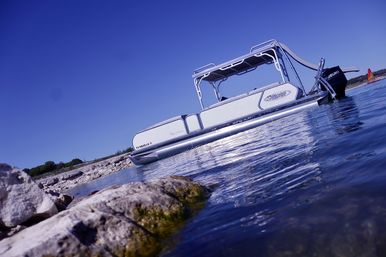 White pontoon boat moored near a rocky lakeshore with calm blue water reflecting sunlight and a distant red sailboat under a clear sky