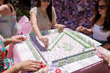 Friends playing mahjong at a sunny floral garden party — pastel green tiles on a pink, patterned table with a flower wall backdrop.