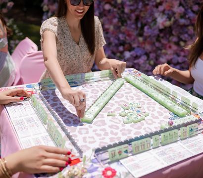 Friends playing mahjong at a sunny floral garden party — pastel green tiles on a pink, patterned table with a flower wall backdrop.