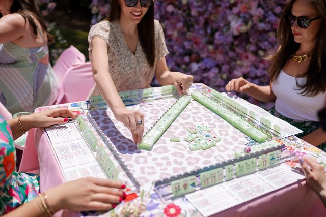 Friends playing mahjong at a sunny floral garden party — pastel green tiles on a pink, patterned table with a flower wall backdrop.