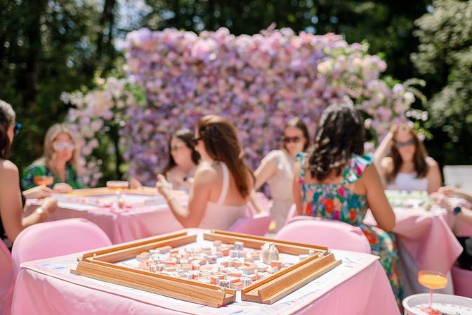 Outdoor spring garden party with friends playing mahjong at pink tables in front of a pastel flower wall backdrop