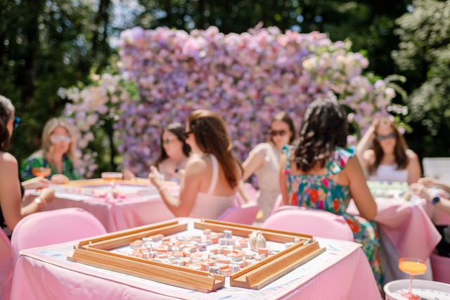 Outdoor spring garden party with friends playing mahjong at pink tables in front of a pastel flower wall backdrop