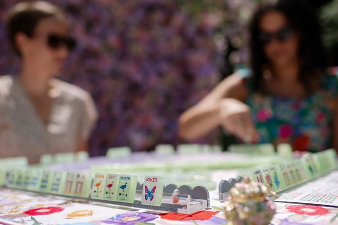 Close-up of Mahjong tiles on racks with a colorful Joker butterfly tile in focus, two blurred players reaching across a floral tablecloth in a sunlit outdoor game.