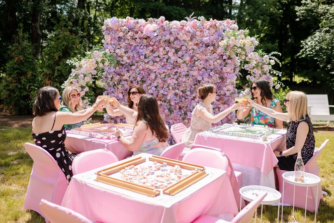 Group of women cheering with cocktails at a pink-themed outdoor garden party, seated around pink-covered tables with tabletop games, against a large pastel flower wall backdrop and leafy trees.