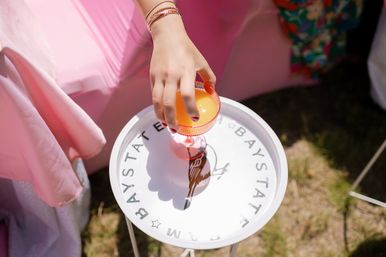 Close-up of a hand with bracelets holding a pink-rimmed coupe glass of orange cocktail on a white round tray with bird motif at an outdoor sunny picnic with pastel pink decor