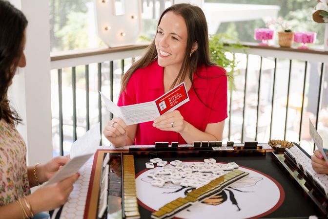Smiling woman in a red blouse holding a scorecard while friends play mahjong around a sunlit balcony game table with tiles, racks and potted plants — casual patio game night