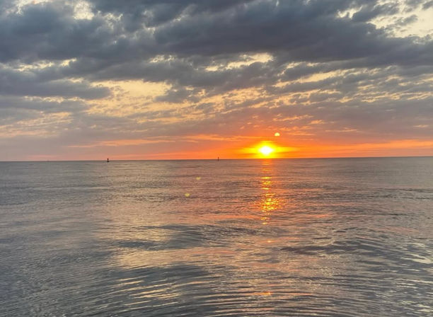 Vibrant golden sunset over a calm ocean, sun casting a sparkling reflection across rippling water beneath a dramatic, cloud-streaked sky on the horizon.
