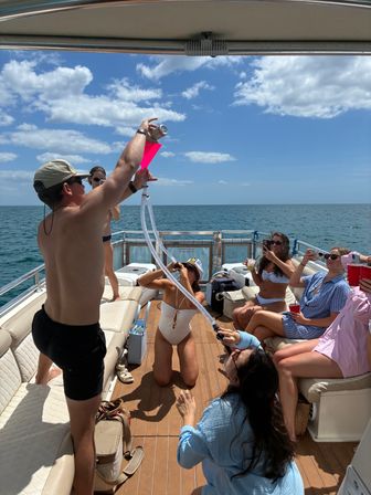 Group of friends on a sunny boat party at sea, a woman kneeling using a long drinking tube and pink funnel while others cheer, hold drinks and take photos under blue sky and scattered clouds.