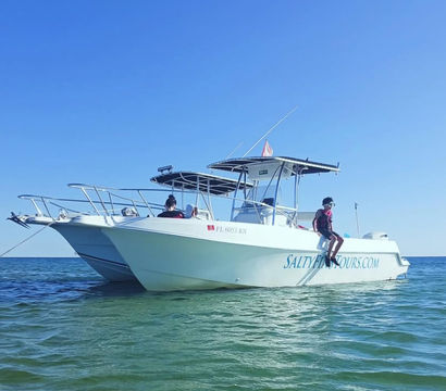 White center-console motorboat anchored in shallow turquoise coastal water under a clear blue sky, with two people on board and one person sitting on the stern with legs dangling for a relaxed boat-tour vibe.