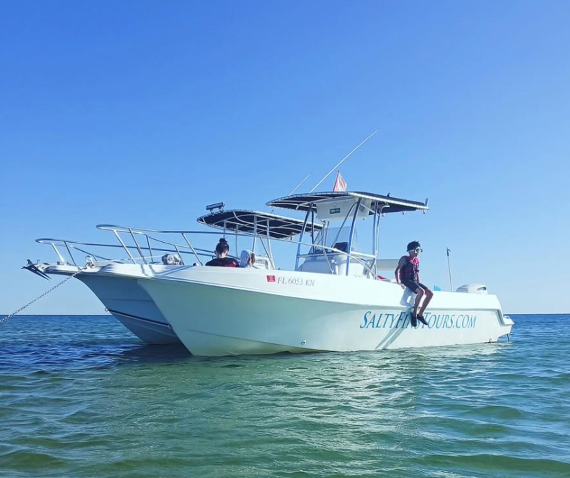 White center-console motorboat anchored in shallow turquoise coastal water under a clear blue sky, with two people on board and one person sitting on the stern with legs dangling for a relaxed boat-tour vibe.