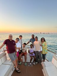 Eight people smiling on a sunset boat cruise over calm ocean waters, wearing casual summer clothes and sunglasses, with a distant coastal skyline and pastel sky.