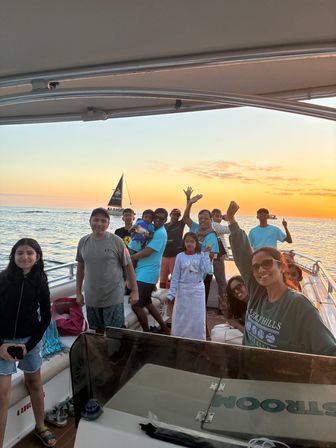 Group of friends and family enjoying a sunset cruise on a motorboat, waving and smiling with a sailboat on calm ocean waters under a golden sky