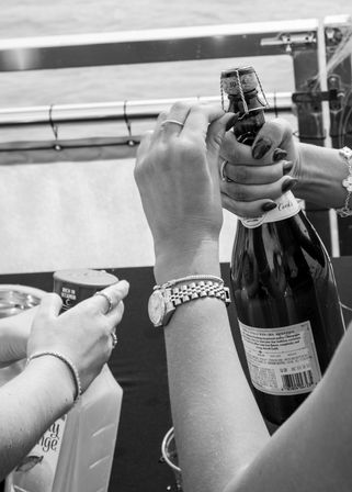 Close-up of hands opening a champagne bottle on a boat deck next to an orange juice carton, wrists with watch and bracelets — ready for mimosas.