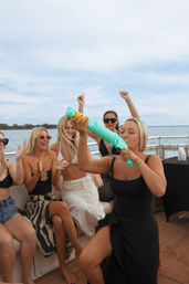 Group of friends partying on a boat over open water, woman in black takes a drink from a turquoise novelty shooter while others cheer and clap under a cloudy sky