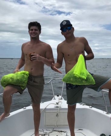 Two shirtless men posing on the bow of a boat at sea, each holding a neon green mesh bag of harvested shellfish under a cloudy sky.