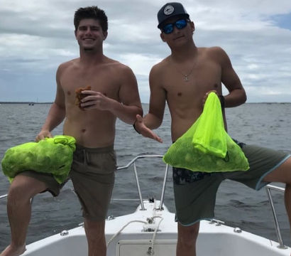 Two shirtless men posing on the bow of a boat at sea, each holding a neon green mesh bag of harvested shellfish under a cloudy sky.