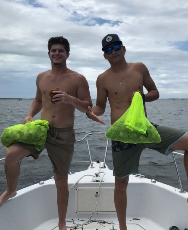 Two shirtless men posing on the bow of a boat at sea, each holding a neon green mesh bag of harvested shellfish under a cloudy sky.