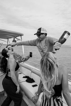 Boat party on coastal waters — friends on a pontoon boat as a man in a cap pours champagne into two women’s mouths, sunglasses and swimsuits, open ocean and cloudy sky in the background.