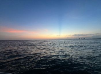Sunset over the open ocean with gentle waves, soft pastel pink and blue sky, and a distant coastal city skyline on the horizon.