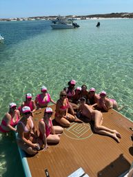 Group of friends in pink swimsuits and matching caps lounging on a floating dock over clear turquoise shallow water near a sandy sandbar with anchored boats and people in the background