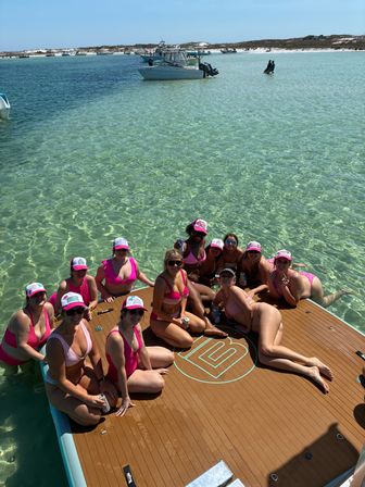 Group of friends in pink swimsuits and matching caps lounging on a floating dock over clear turquoise shallow water near a sandy sandbar with anchored boats and people in the background