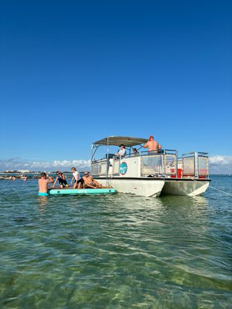 Group enjoying a sunny day on a party-style pontoon boat with people on a paddleboard in shallow turquoise water under a clear blue sky