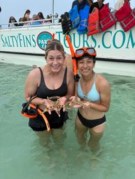 Two smiling women standing in shallow turquoise water holding starfish during a coastal snorkeling boat tour, wearing snorkel gear with life jackets hanging on the boat behind them.
