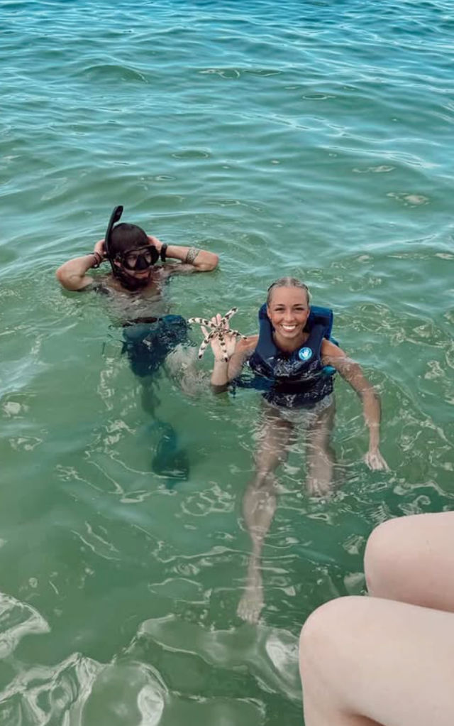 Two people snorkeling in clear turquoise sea, one in a life vest smiling while holding a spotted starfish and the other adjusting a snorkel mask near a boat