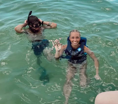 Two people snorkeling in clear turquoise sea, one in a life vest smiling while holding a spotted starfish and the other adjusting a snorkel mask near a boat