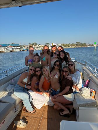 Group of friends in swimsuits posing on a boat deck at a sunny coastal marina with docks and boats in the background