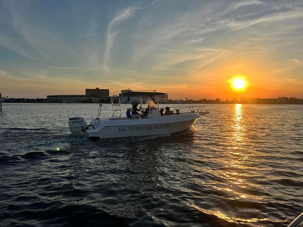 White motorboat with passengers cruising across a bay at golden sunset, sun reflecting on rippled water and a low waterfront skyline on the horizon.