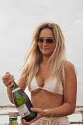 Blonde woman in a white bikini and sunglasses opening a champagne bottle on a boat off a sandy beach, summer coastal celebration vibe